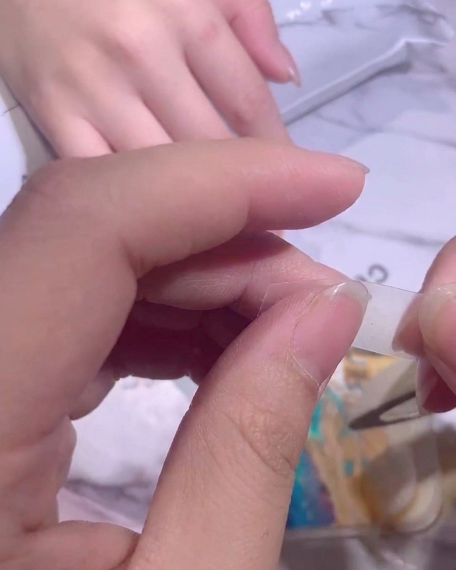 Des doigts plaçant soigneusement une fine bande de ruban adhésif transparent sur un ongle pour bloquer la base.