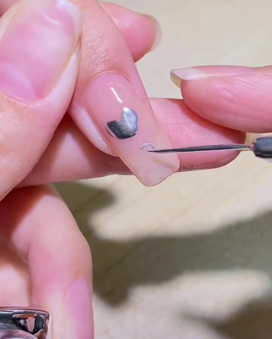 Gros plan dun pinceau liner dessinant le contour dun cœur argenté sur un ongle transparent.