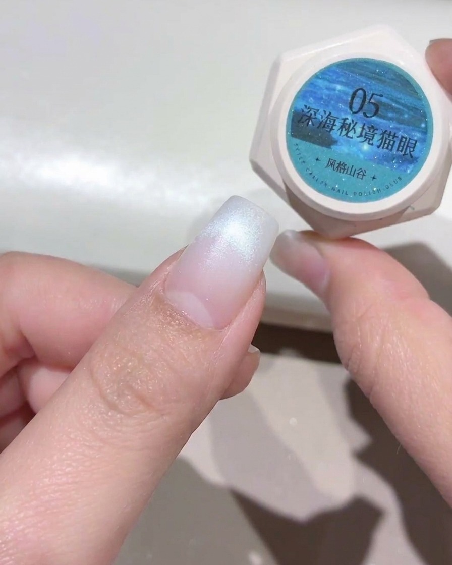 Une main tenant un petit pot de gel cat eye bleu canard à côté dun ongle préparé avec une pointe blanche.