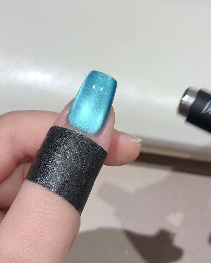 Un petit aimant sphérique tenu près du bord dun ongle bleu, concentrant les reflets pour un effet perle 3D lumineux.
