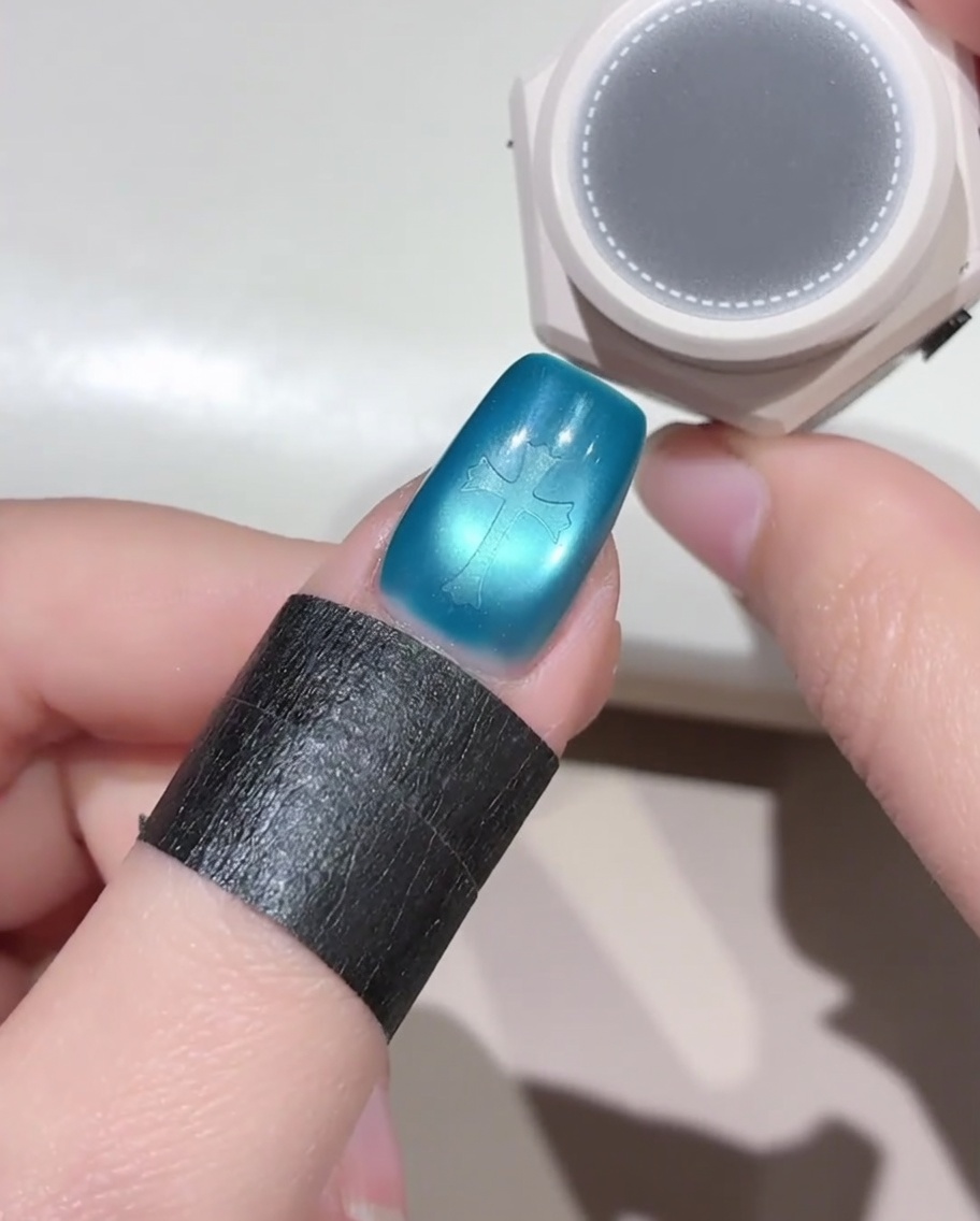 Un petit outil en éponge ronde planant au-dessus dun ongle bleu orné dun autocollant en forme de croix, prêt à appliquer du vernis gel noir.