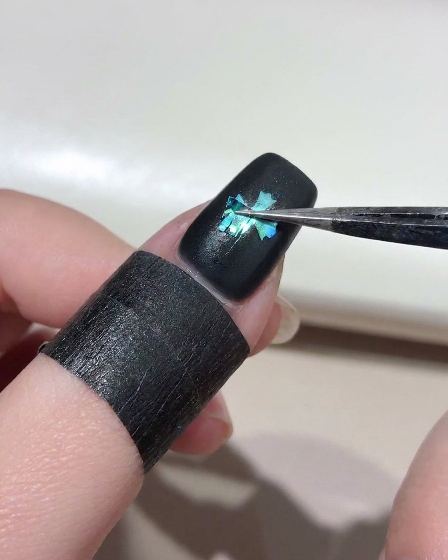 Une pince à épiler soulevant délicatement un pochoir en forme de croix dun ongle peint en noir, révélant une croix cat eye bleu vif en dessous.