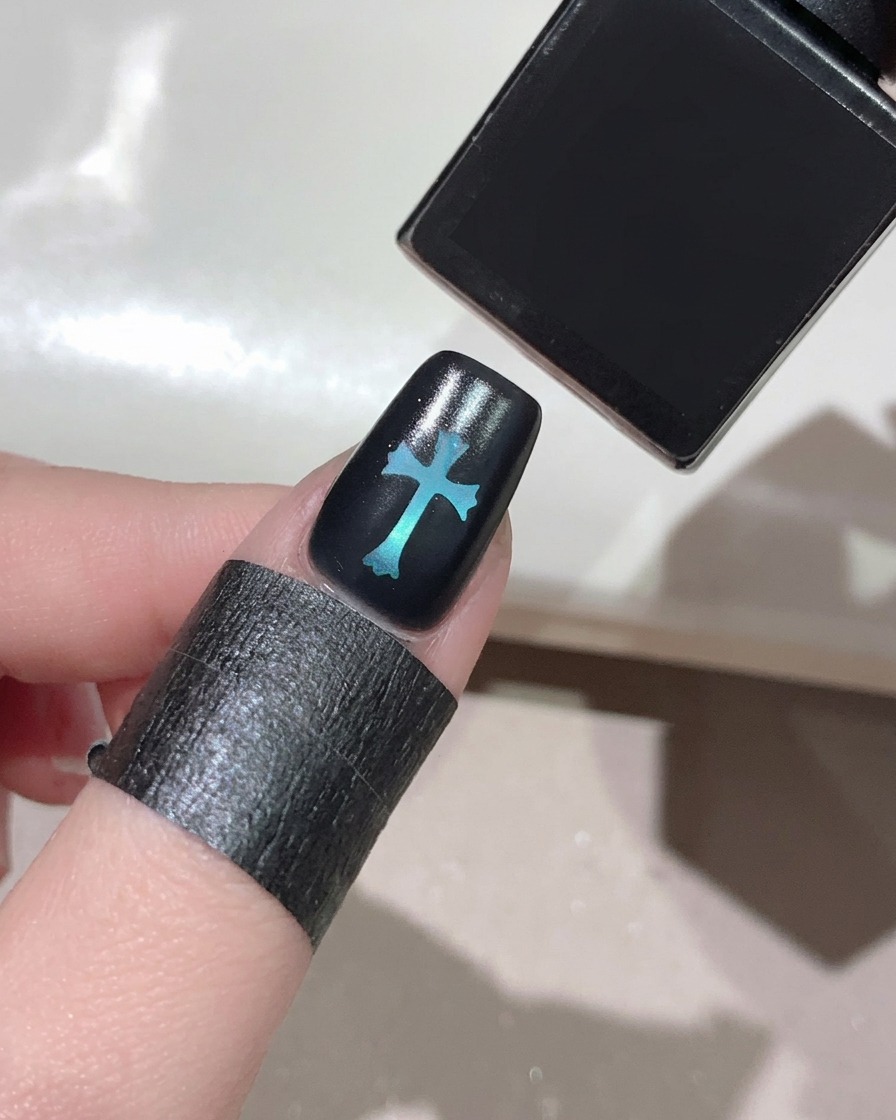 Un pinceau appliquant un top coat transparent et brillant sur un ongle noir avec un motif de croix bleue lumineuse au centre.