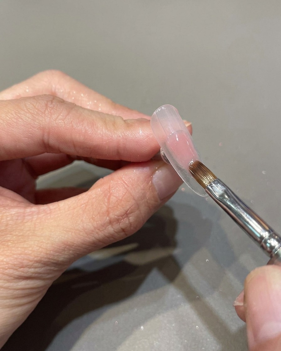 Un pinceau de manucure professionnel appliquant un vernis gel rose translucide sur un ongle préparé.