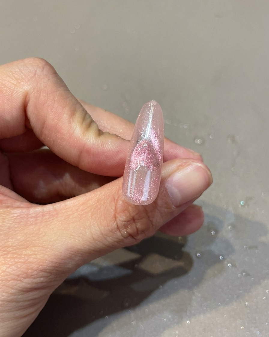 Gros plan sur un ongle rose avec un design de cœur cat-eye, montrant un aimant utilisé pour manipuler leffet scintillant.
