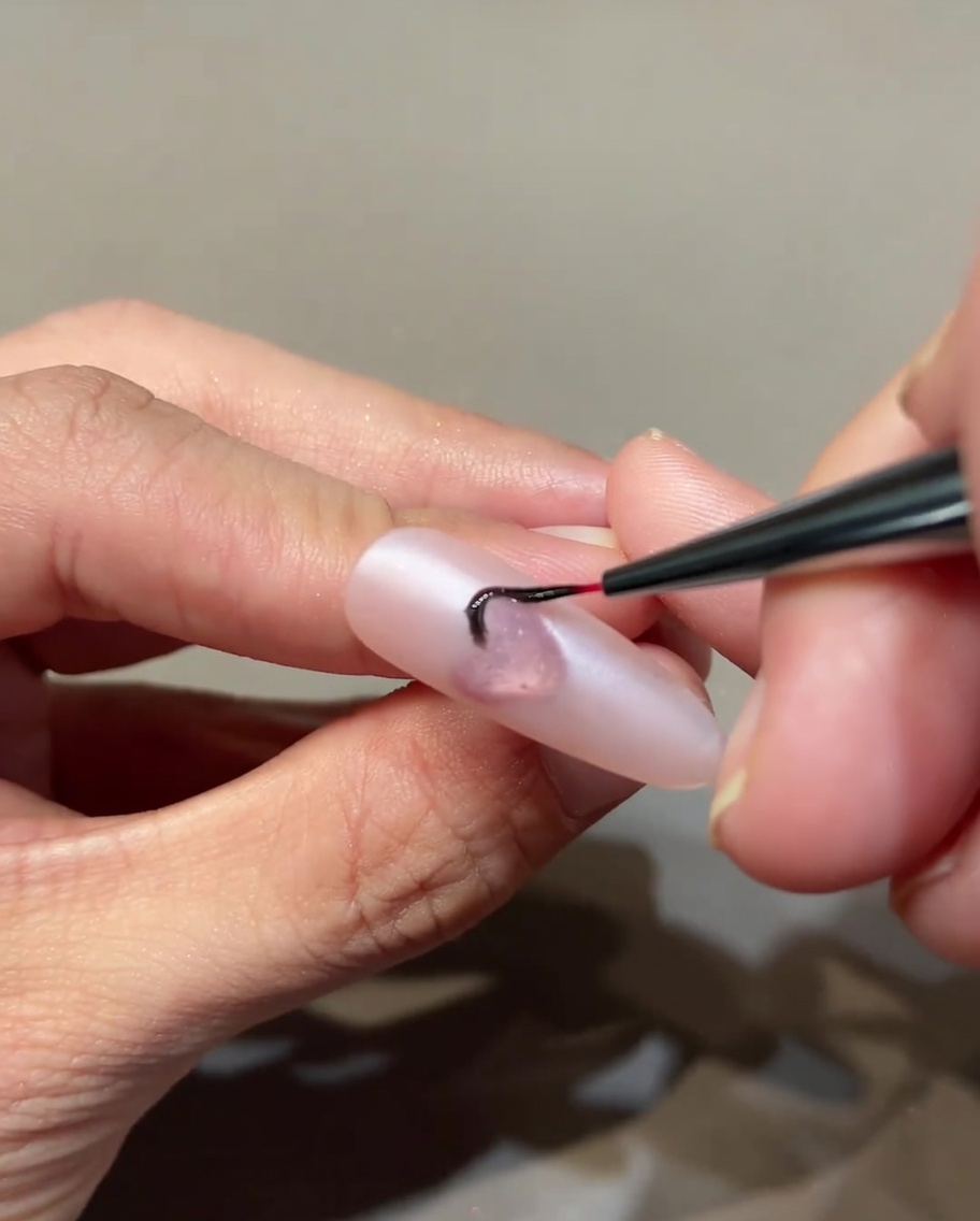 Un pinceau nail art fin dessinant des flammes noires fines sur une base rose autour dun cœur central.