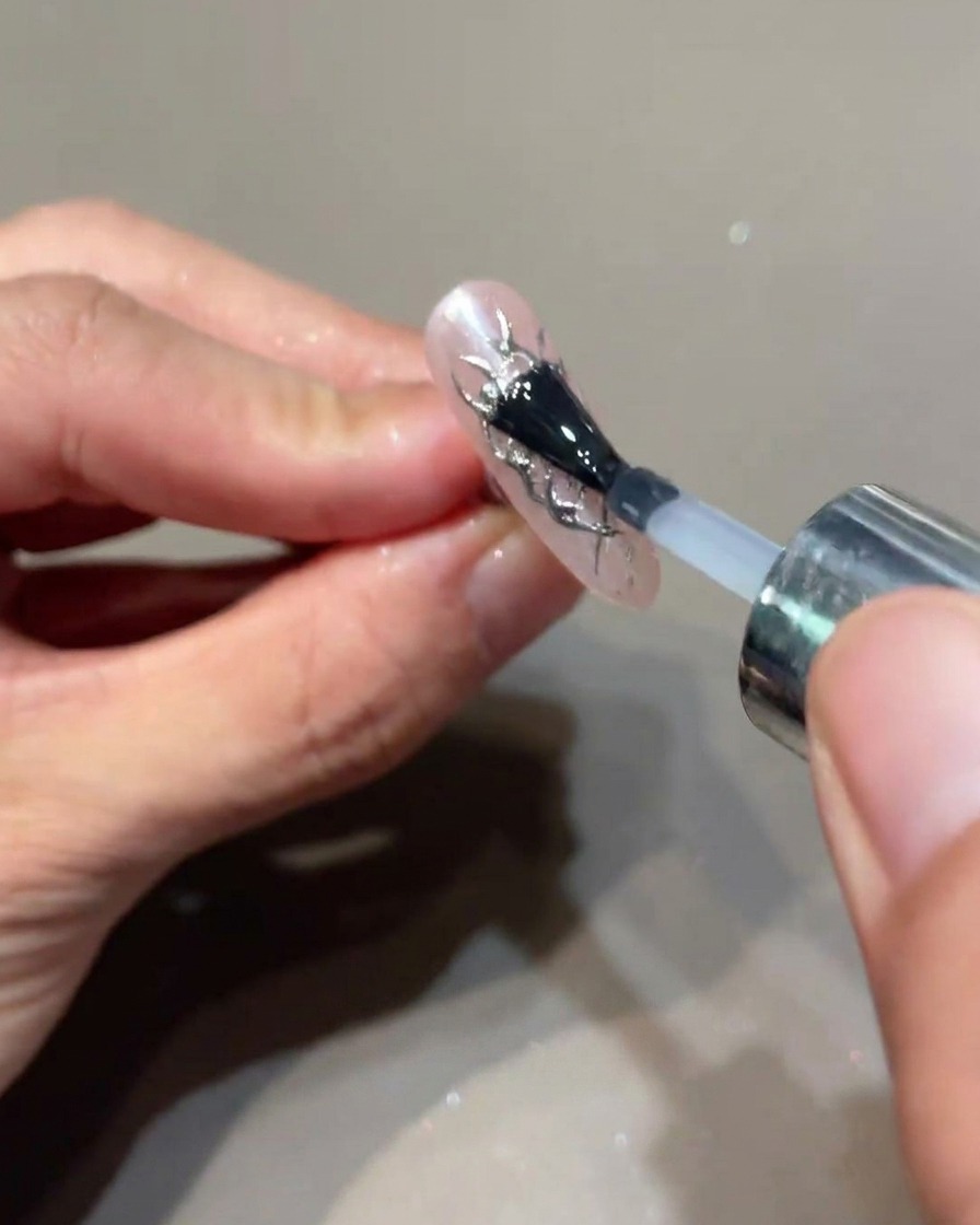 Un pinceau appliquant une dernière couche de top coat transparent sur un ongle décoré de flammes argentées.