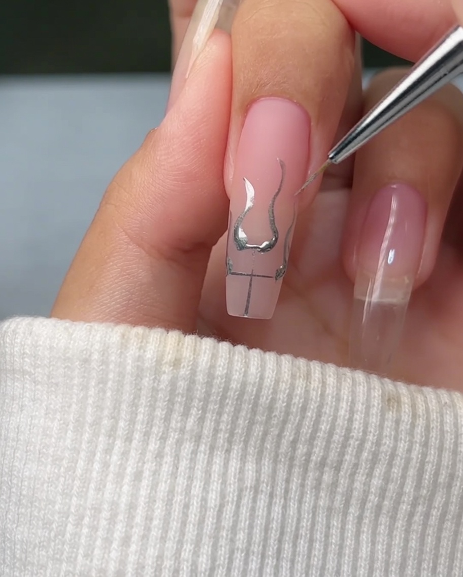 Un pinceau fin remplissant la base des contours de flammes sur un ongle avec un gel argent métallique brillant et réfléchissant.