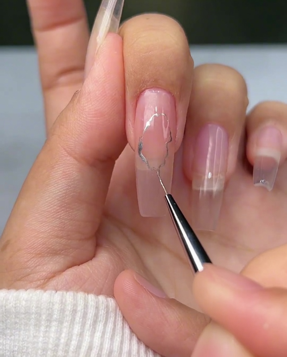 Un pinceau liner fin dessinant des lignes argentées irrégulières et ondulées sur un ongle.
