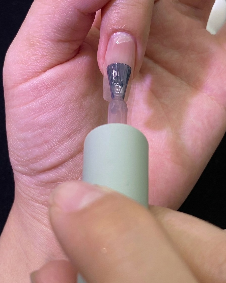 Gros plan dun pinceau appliquant un vernis semi-permanent nude translucide sur un ongle naturel.