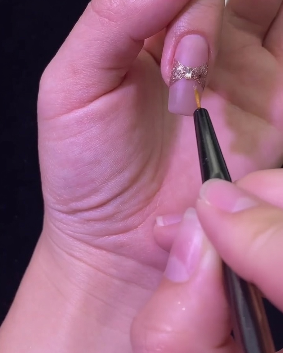 Un pinceau liner appliquant un top coat brillant pour sceller le nœud en paillettes or rose et le strass central.
