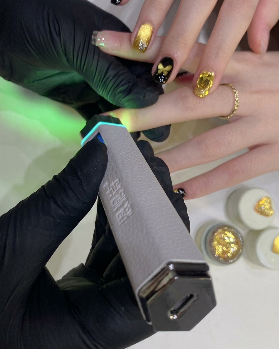 Une main gantée tenant une lampe UV/LED portative au-dessus dun ongle peint au vernis gel noir.
