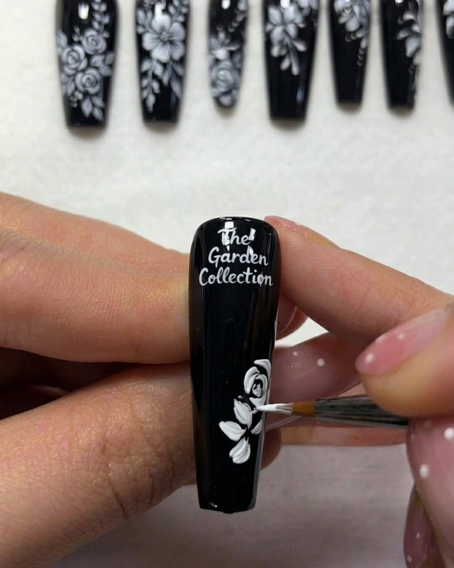 Vue rapprochée dune styliste ajoutant des lignes blanches fines pour représenter des tiges partant du feuillage sur un ongle peint en noir.