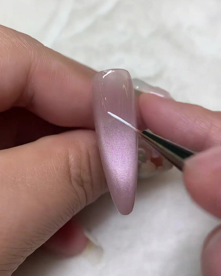 Gros plan dun pinceau liner appliquant un fin trait blanc vertical au centre dun ongle en gel cat-eye mauve.