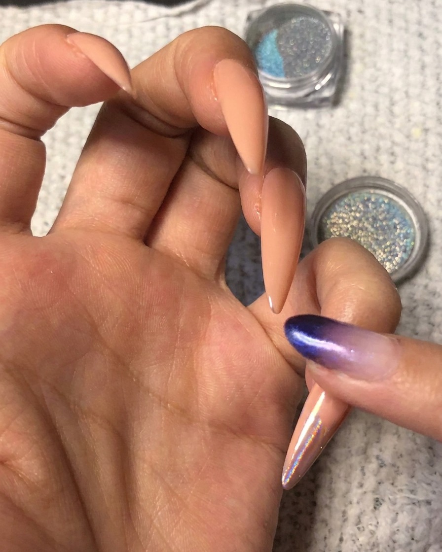 Un doigt pressant de la poudre chrome holographique bleu-violet vibrant sur le bout dun ongle nude en forme damande.