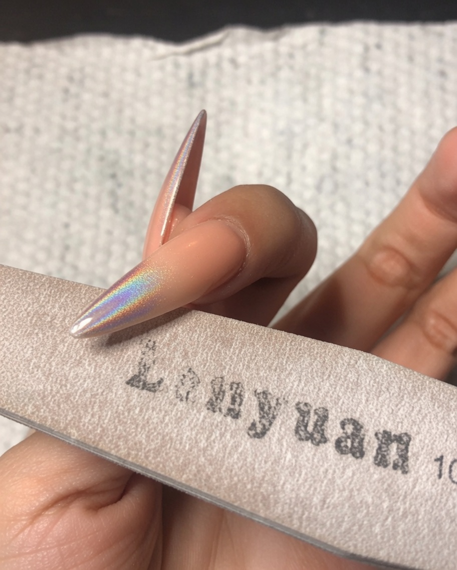 Une main utilisant une lime éponge fine pour polir délicatement le bord libre dun ongle stiletto décoré avec de la poudre chrome holographique.
