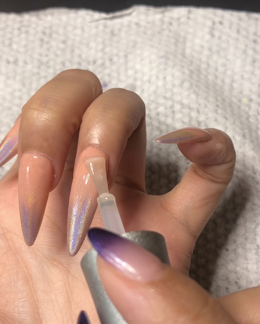 Un pinceau à ongles appliquant un top coat ultra-brillant sur lannulaire, scellant le nail art ombré holographique.