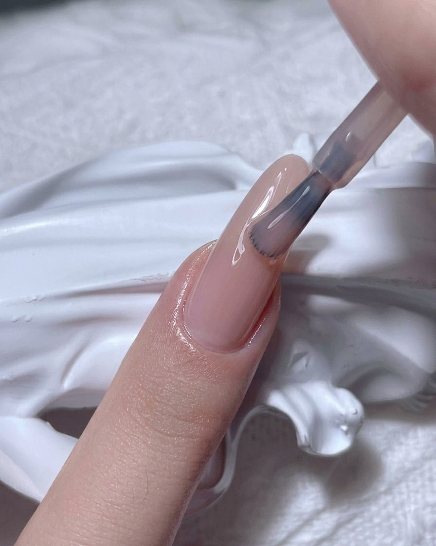 Un pinceau appliquant un vernis gel nude transparent sur un ongle naturel.