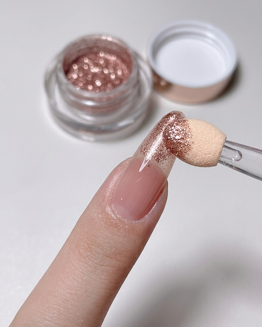 Un applicateur éponge frottant de la poudre or rose sur la pointe dun ongle nude.