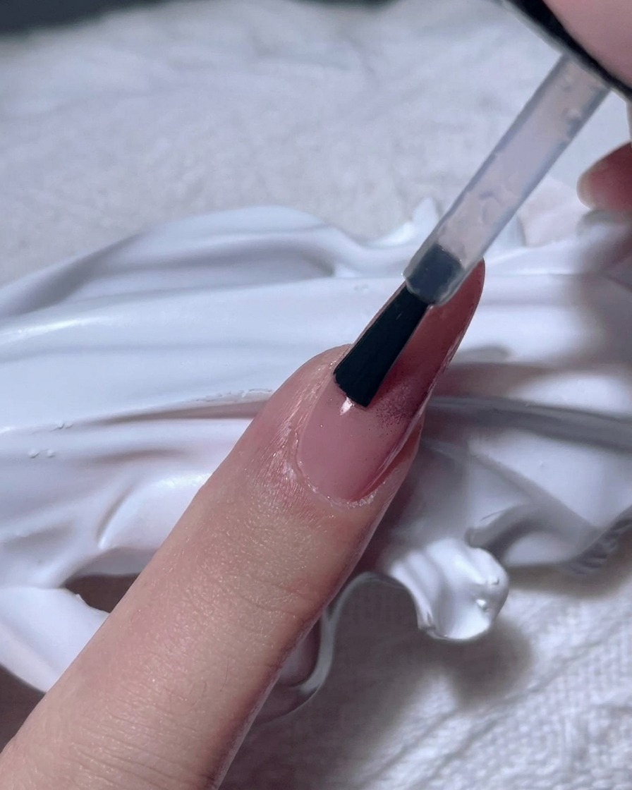 Un pinceau appliquant un gel transparent épais sur un ongle en chrome or rose.