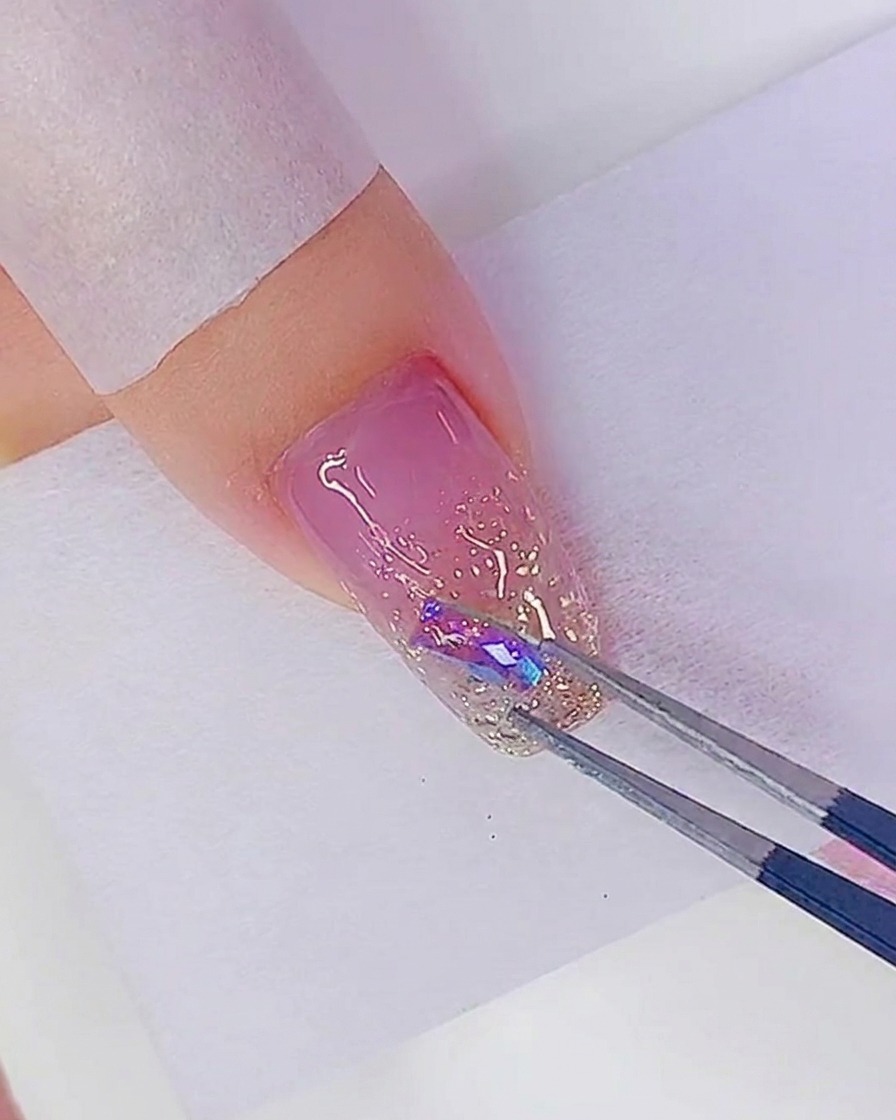 Des pincettes plaçant un strass iridescent sur un point de colle sur un ongle pailleté.
