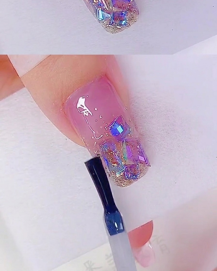 Un pinceau à ongles appliquant un top coat transparent sur un ongle décoré de paillettes or rose et de strass iridescents.