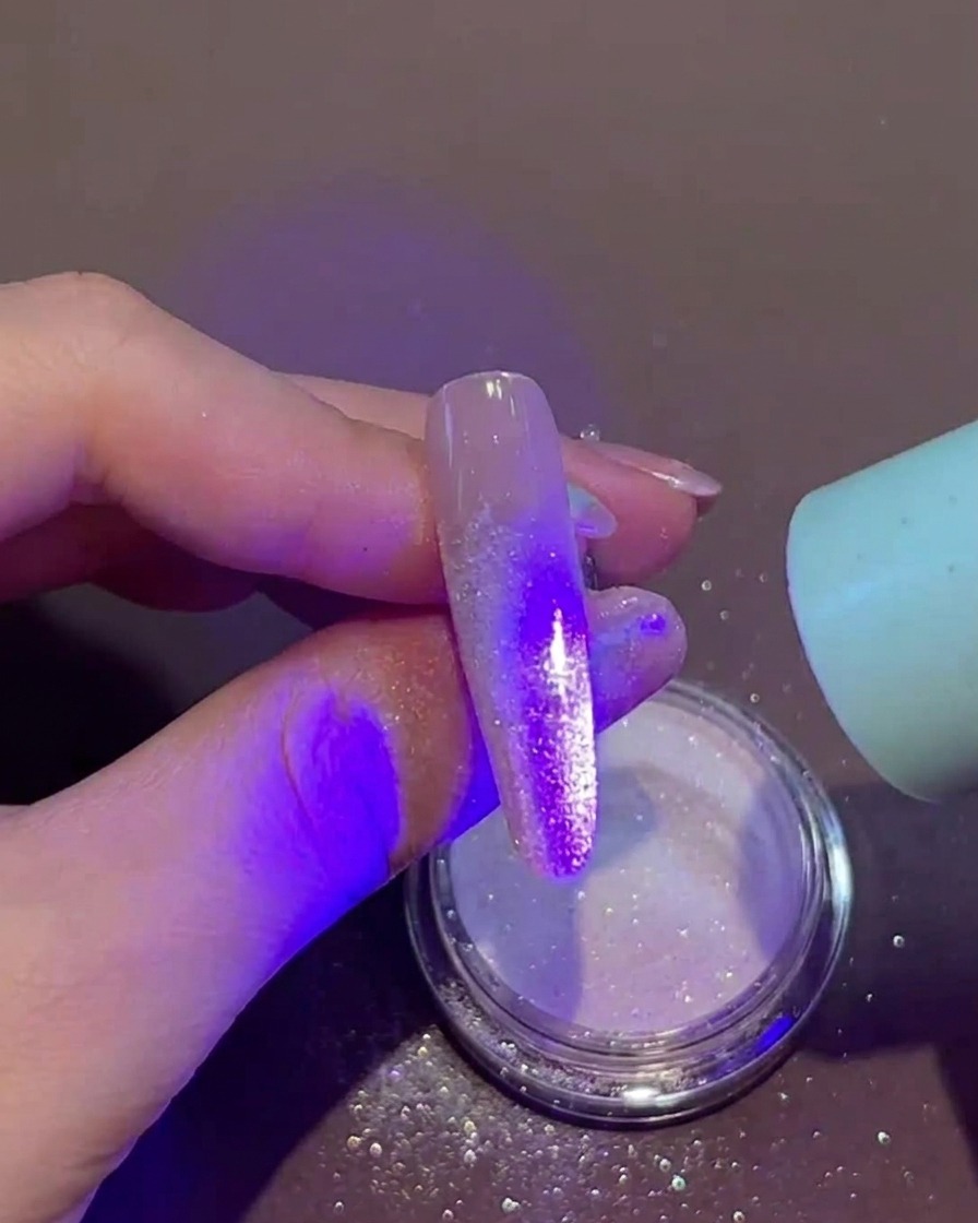 Une main tenant une petite lampe UV/LED portable directement au-dessus dun ongle avec du gel humide et de la poudre libre sur la pointe.
