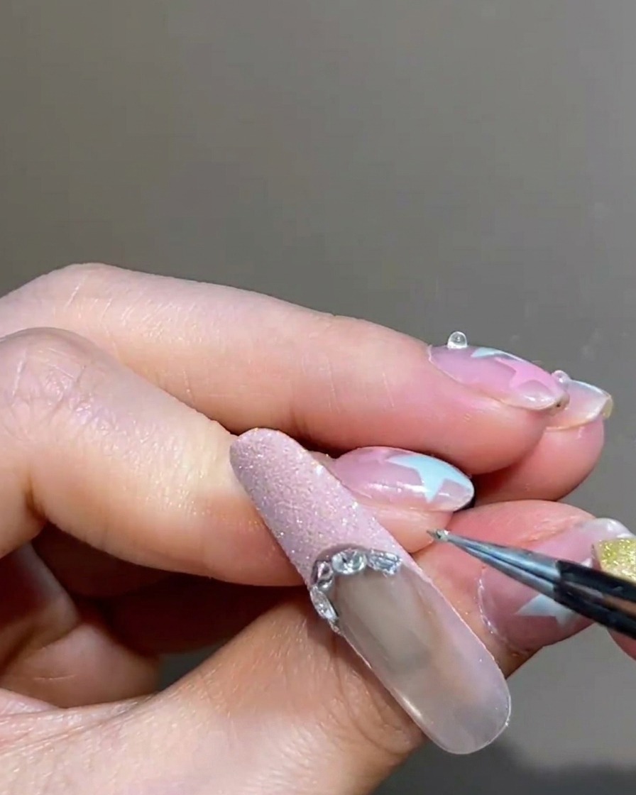 Gros plan dune pince à épiler plaçant une petite perle métallique argentée le long de la délimitation dune French rose sur un ongle manucuré.