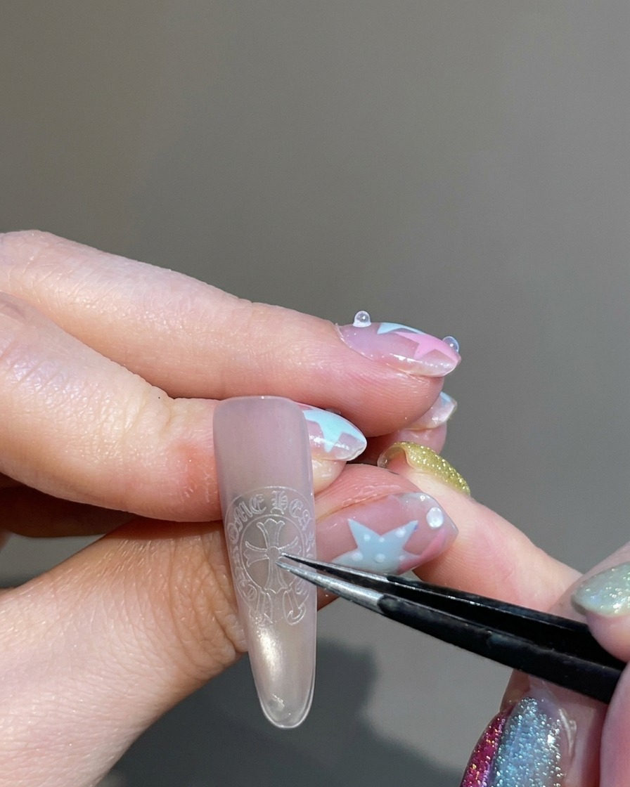 Une pince à épiler tenant et plaçant un autocollant en forme de croix au centre dun ongle.