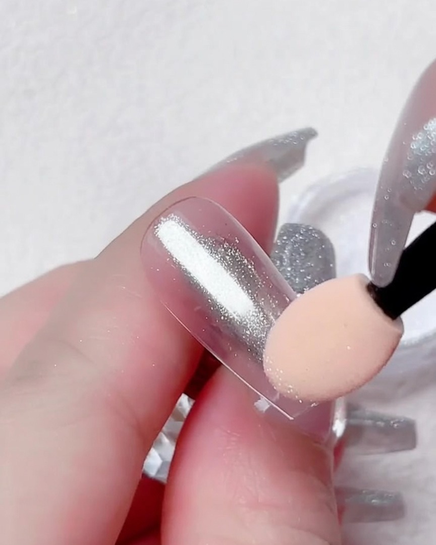 Gros plan dun doigt tenant une capsule transparente pendant quun applicateur mousse dépose de la poudre chrome argentée.