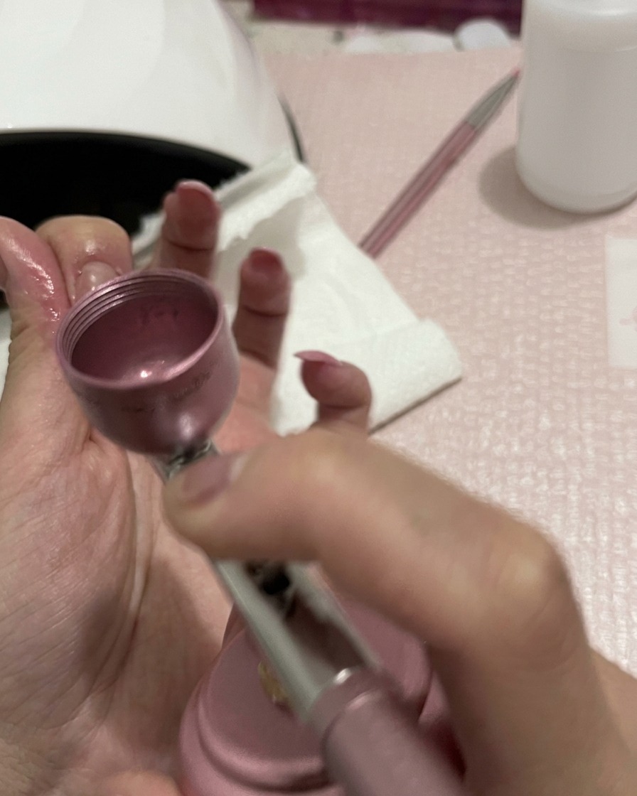 Vue rapprochée de mains tenant un aérographe, avec la buse concentrée sur le mélange de la couleur pêche sur les ongles.