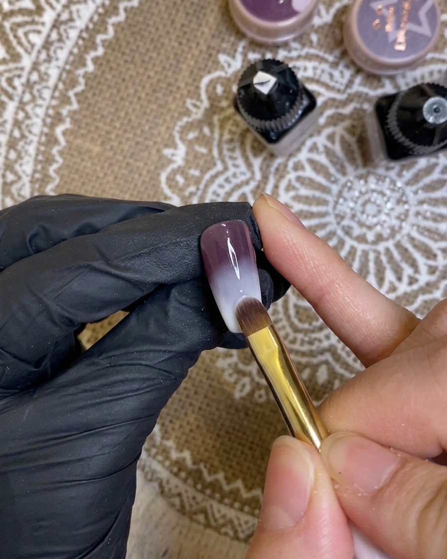 Gros plan sur un pinceau appliquant un gel violet riche et foncé sur le bord dun ongle manucuré.