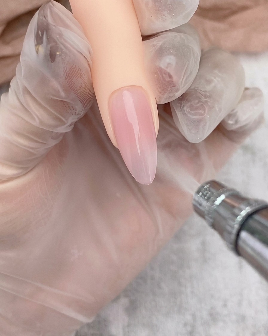 Un aérographe appliquant un dégradé blanc doux sur le bord dun ongle avec base gel transparente.