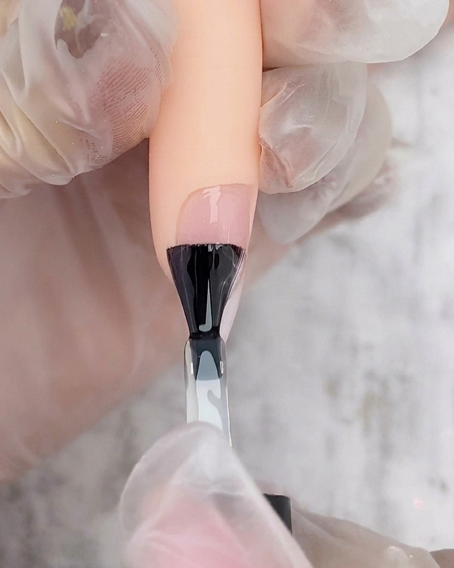 Un pinceau de vernis noir appliquant une couche de top coat gel transparent sur un ongle ombré blanc et rose.