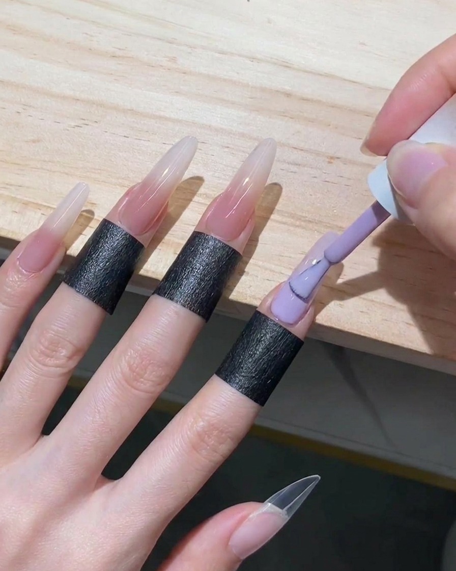 Un pinceau appliquant du gel nude translucide sur la partie inférieure dun ongle stiletto, remontant jusquà rejoindre le bord blanc estompé.