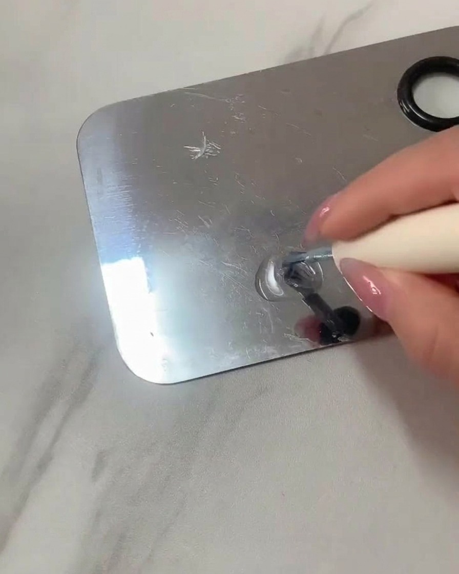 Un pinceau plat mélangeant du gel transparent et du vernis bleu clair sur une palette en acier inoxydable.