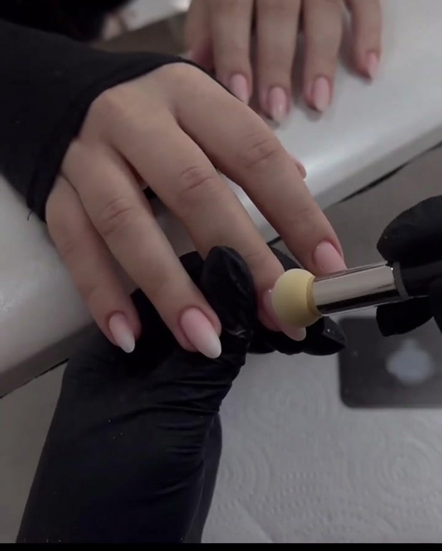 Une main gantée utilisant un applicateur mousse pour estomper la poudre chrome nacrée vers le bas sur un ongle rose nude.