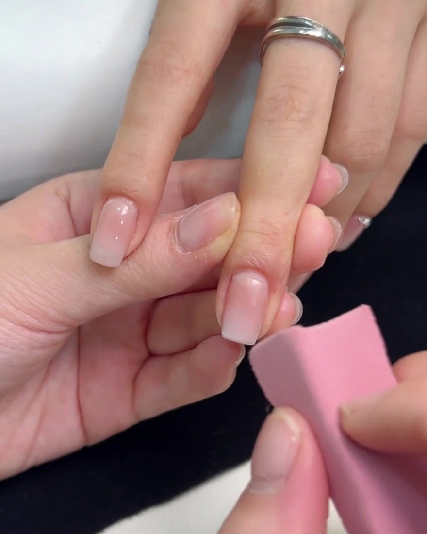 Une éponge rose utilisée pour tapoter du vernis gel blanc sur le bout dun ongle carré, en le fondant vers le bas pour créer un effet ombré.