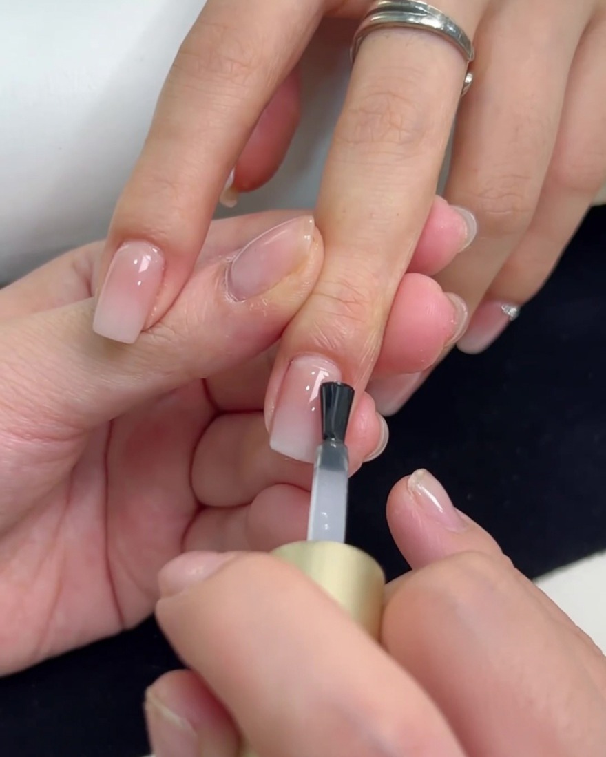 Un pinceau appliquant une couche de finition transparente de renfort sur un ongle au dégradé blanc laiteux.