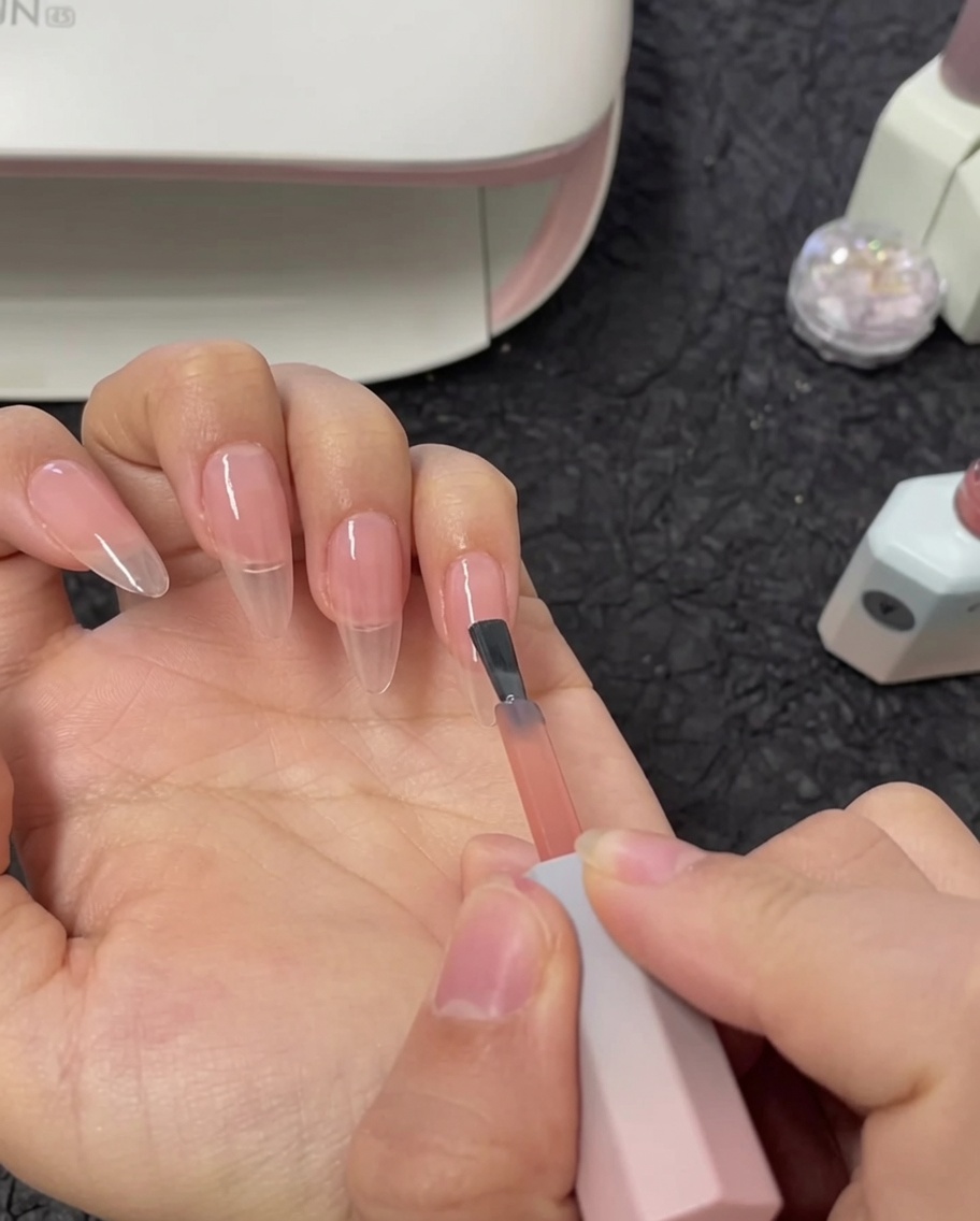 Un pinceau appliquant une seconde couche de vernis gel nude sur un ongle index.