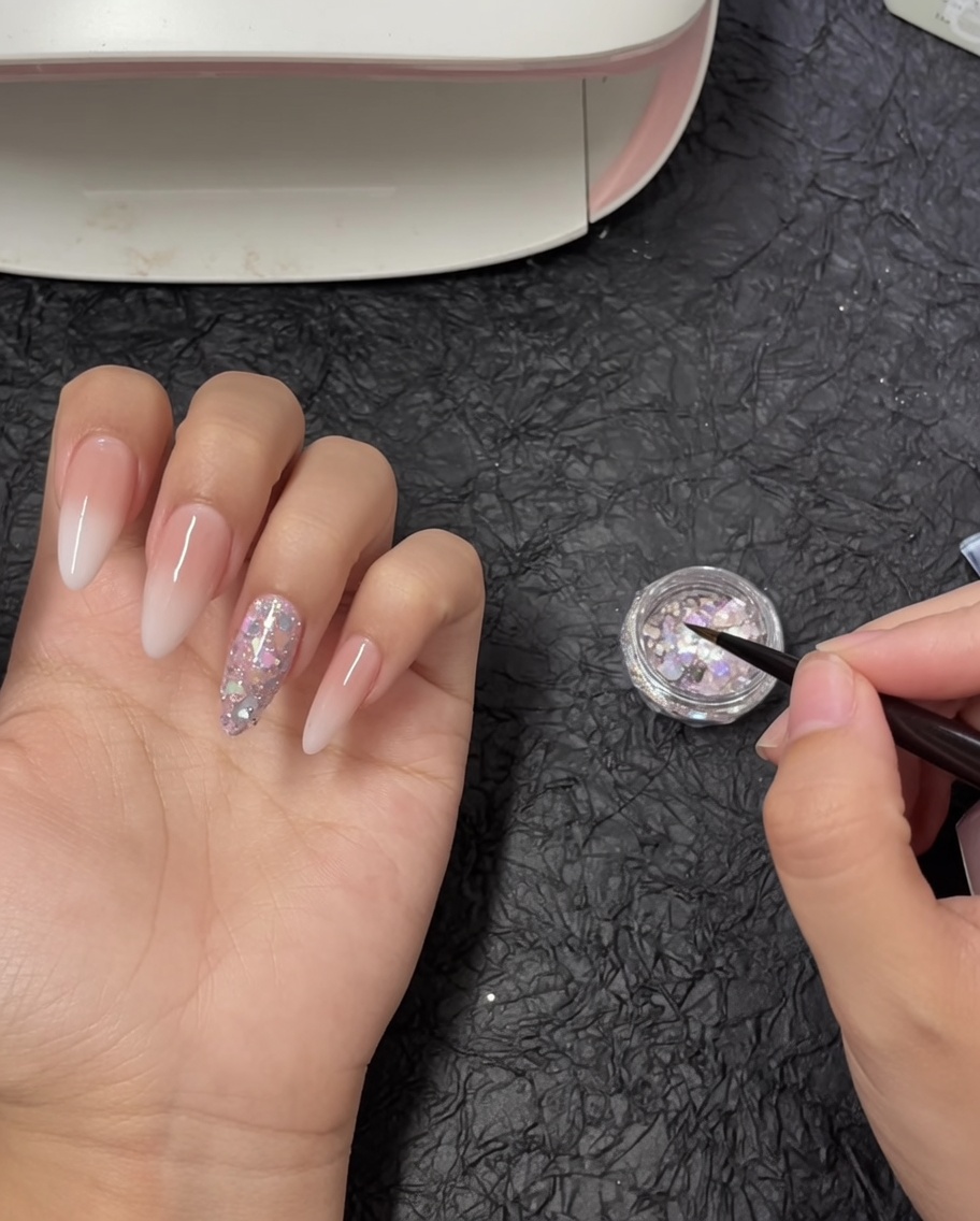 Un pinceau de précision pour nail art plongeant dans un petit pot rempli de flocons de paillettes holographiques épaisses.