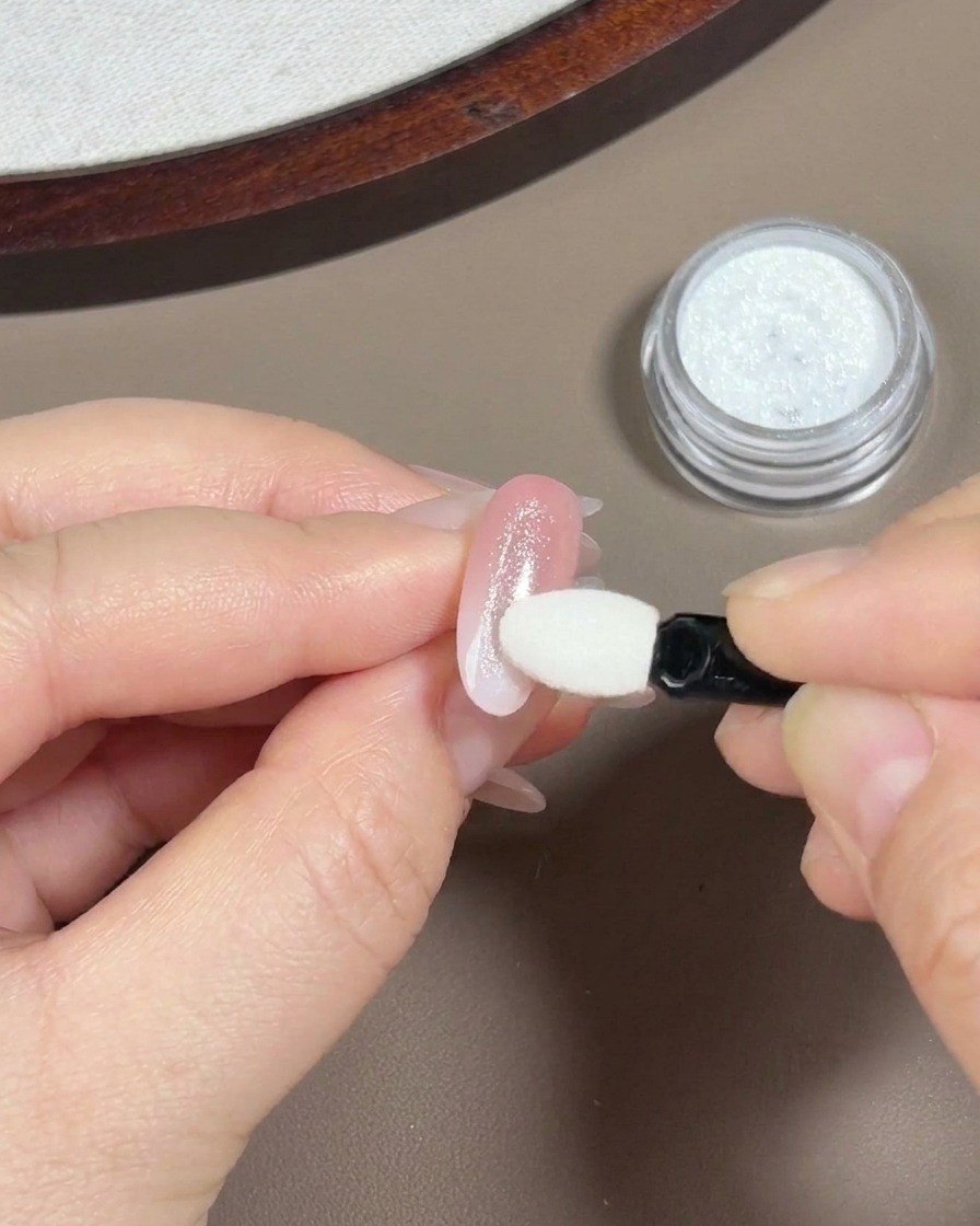 Une main utilisant un petit applicateur mousse pour ombre à paupières pour frotter une poudre aurora irisée sur un ongle au dégradé rose clair.