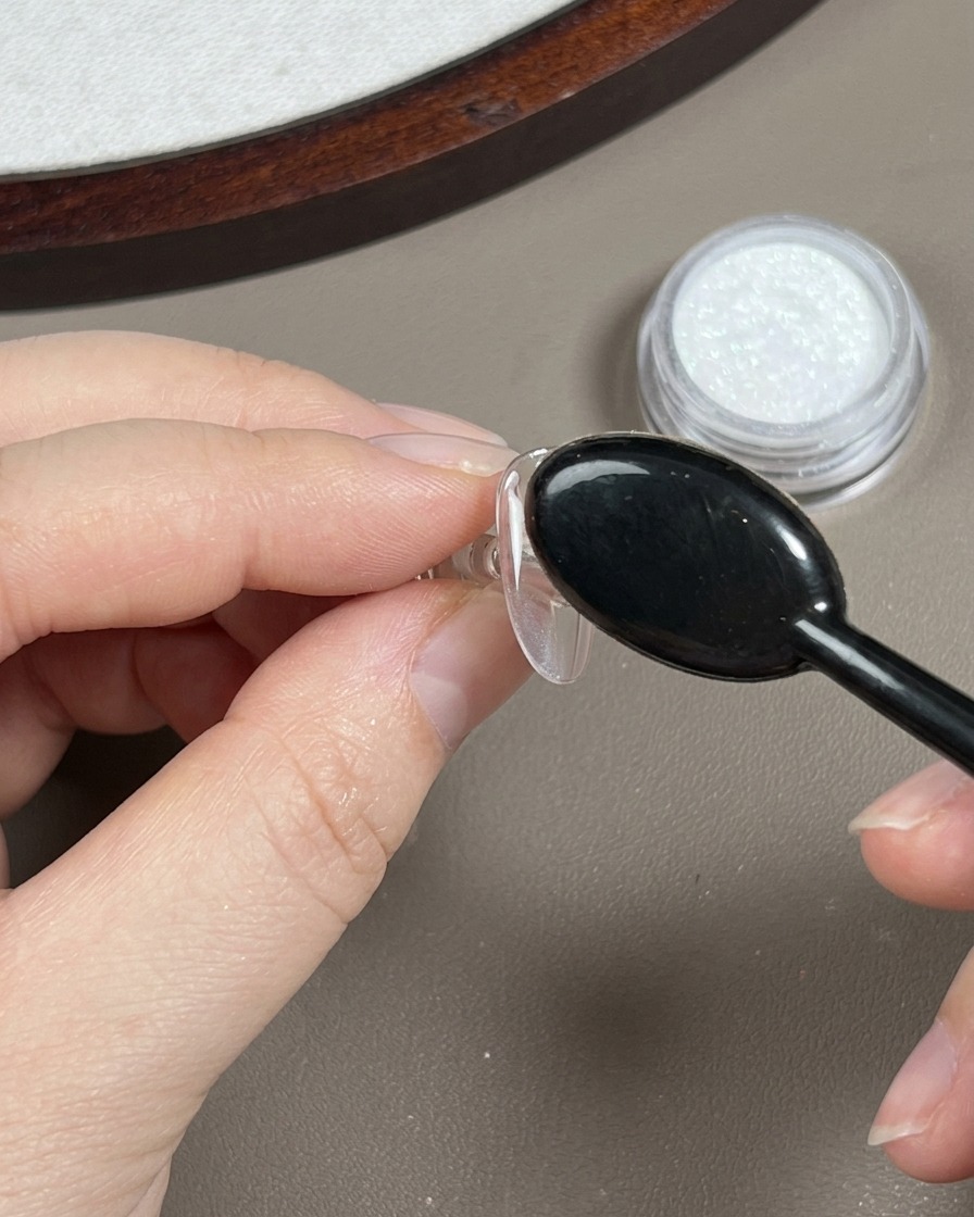 Un pinceau plumeau ovale noir balayant un ongle verni pour enlever la poudre chrome translucide en excès.