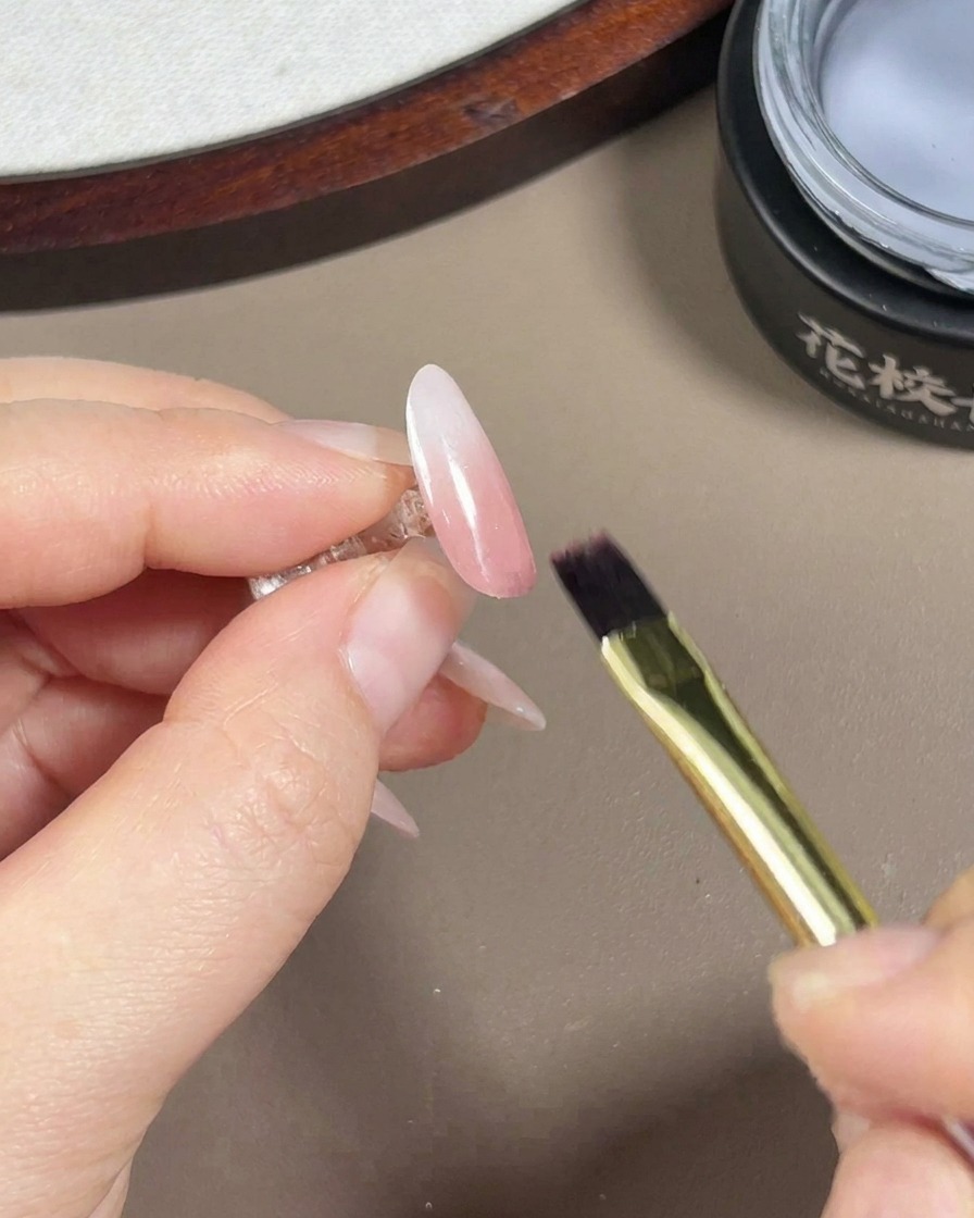 Un pinceau pour gel appliquant une couche de finition transparente et brillante sur un ongle au dégradé rose et blanc avec finition aurora.