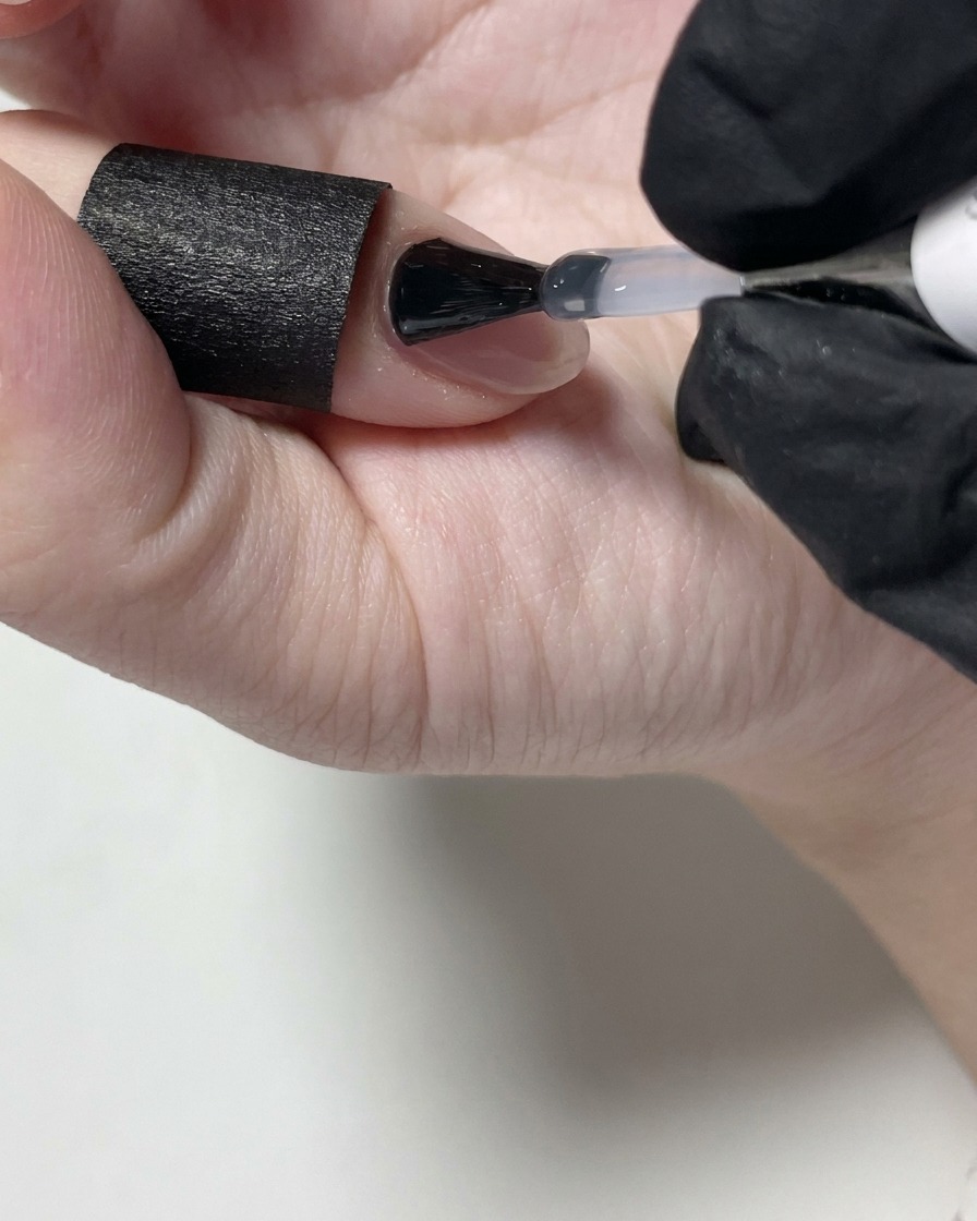 Une styliste ongulaire appliquant un top coat mat transparent sur un ongle en gel nude à laide dun pinceau.