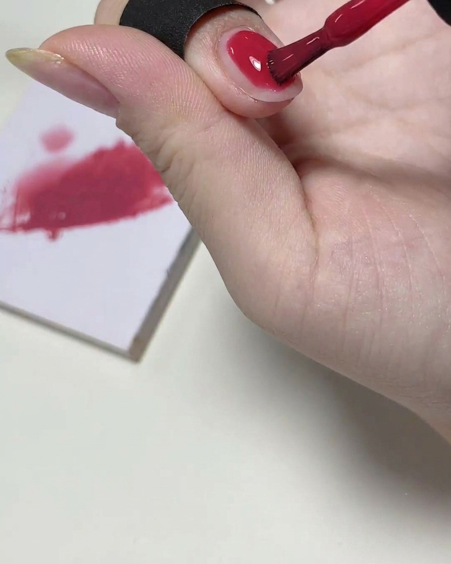 Un pinceau à gel appliquant une couche de vernis rose sur la moitié supérieure de longle, palette de mélange en arrière-plan.