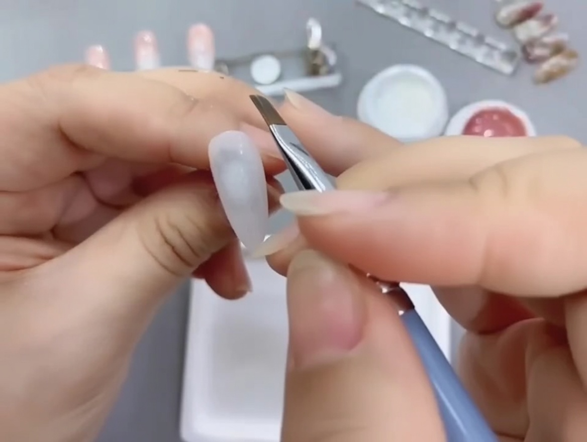 Un pinceau nail art biseauté appliquant du vernis gel nude sur la moitié inférieure dun ongle peint en blanc.