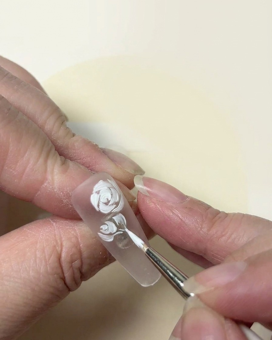 Gros plan pictural sur un design de rose blanche en cours daffinage avec un pinceau à pointe fine sur un ongle transparent.