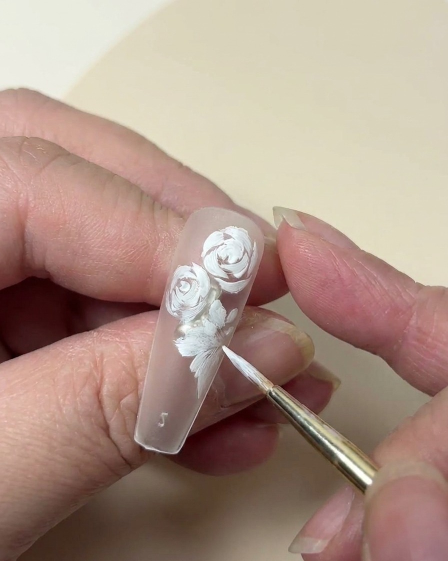 Gros plan sur un ongle montrant une rose peinte en blanc complétée par une petite fleur à cinq pétales et des détails de feuilles.