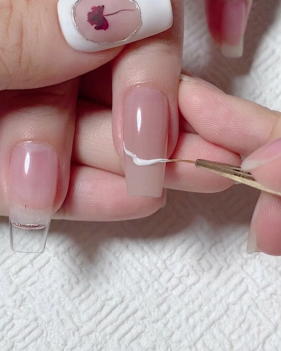 Un pinceau liner dessinant une ligne blanche ondulée au centre dun ongle nude.
